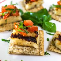 A white plate filled with several crackers topped with Grilled Halloumi cheese, diced tomato, and fresh sweet basil. Fresh sweet basil is in the center.