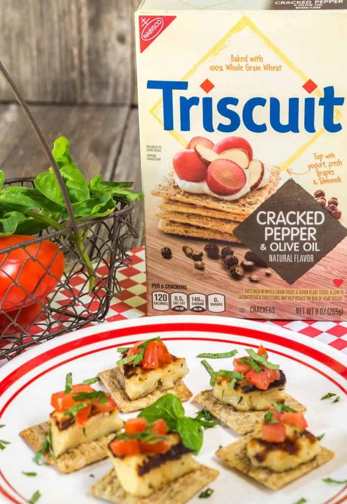 A white plate filled with several crackers topped with Grilled Halloumi cheese, diced tomato, and fresh sweet basil. Fresh sweet basil is in the center. A Triscuit box and a basket of fresh tomatoes and sweet basil sit in the background.