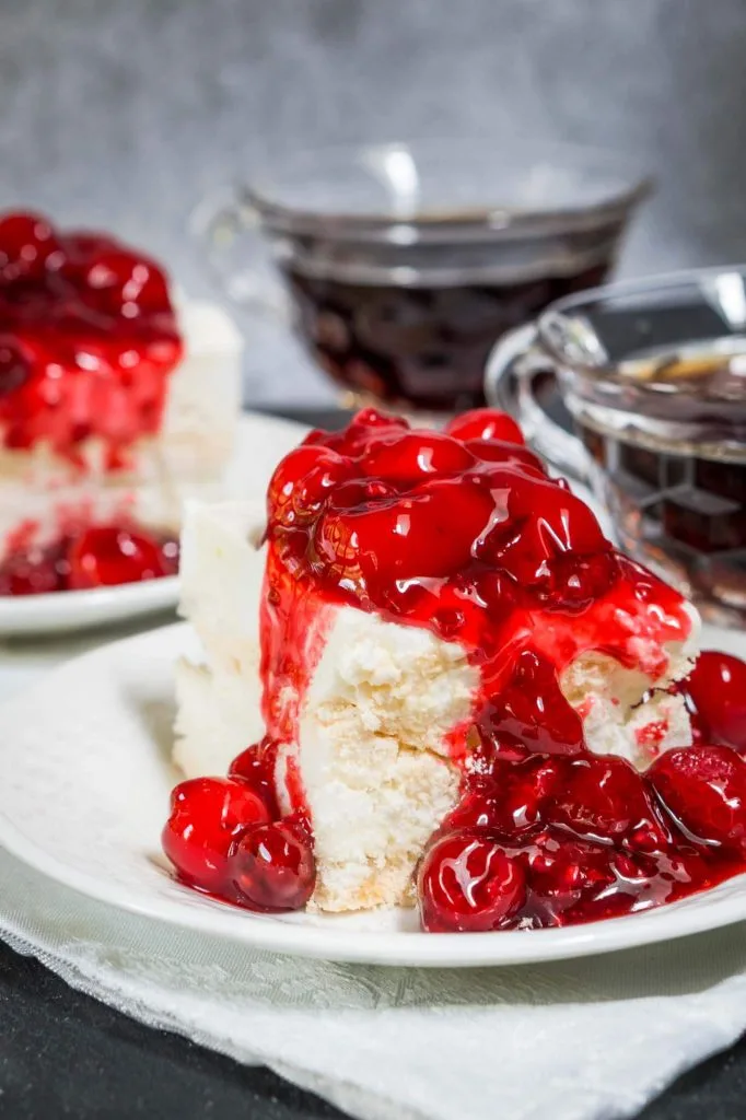 Angled photo of a piece of baked Meringue topped and spilling down the side with red cherries. A second piece and two cups of coffee sit in the background.