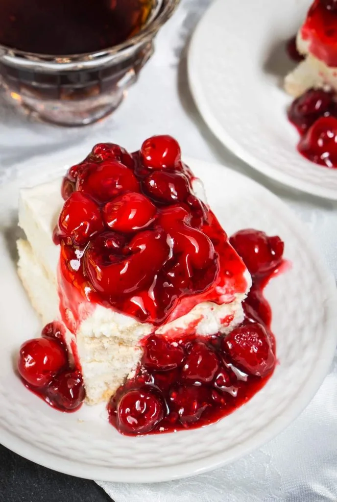A piece of baked Meringue topped and spilling down the side with red cherries. Another plate and a coffee cup sit in the background.