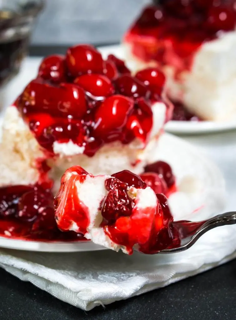 Table view of a piece of baked Meringue topped and spilling down the side with red cherries. A forkful of the dessert hovers in front of the dessert and a second piece appear in the background.