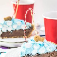 A slice of ice cream pie designed like a swimming pool sitting on a white plate over a tablecloth with colorful circles on it. A crust is filled with chocolate filling topped with blue-hued cool whip and mini marshmallows. A fishing pole with a red striped string is attached to a graham cracker fish. A second slice sits in the background. Red cups sit next to each slice of pie.
