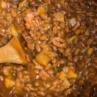 Looking down at a cast iron pot filled with baked beans, apples, shredded pork and jalapeno peppers with a wooden spoon on the side.