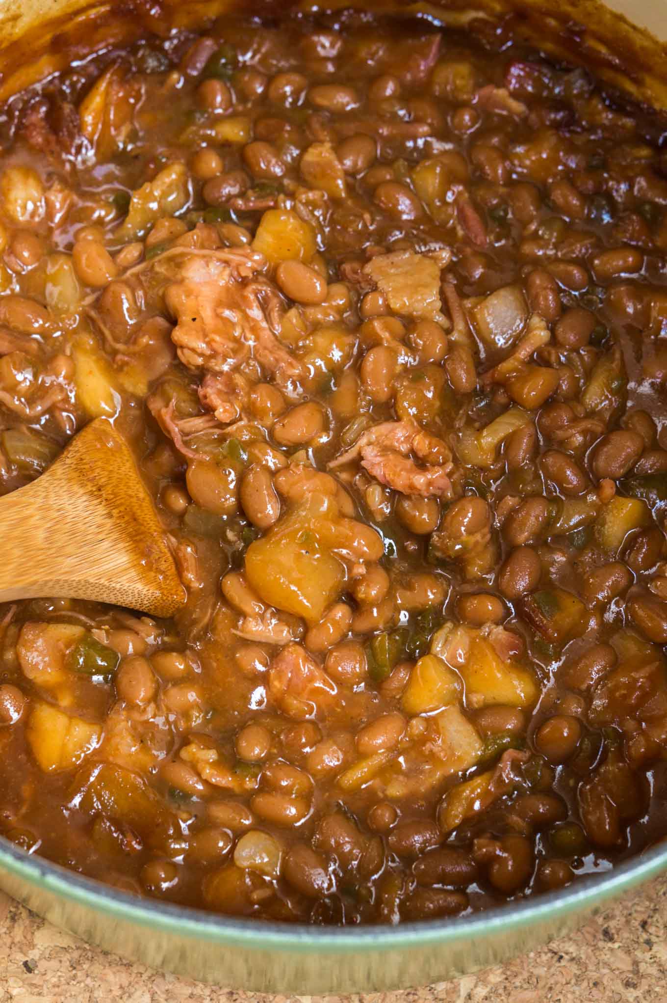 Looking down at a cast iron pot filled with baked beans, apples, shredded pork and jalapeno peppers with a wooden spoon on the side.