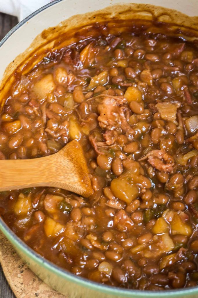 Mirando hacia abajo a una olla de hierro fundido llena de frijoles horneados, manzanas, cerdo rallado y jalapeño pimientos con una cuchara de madera al lado.