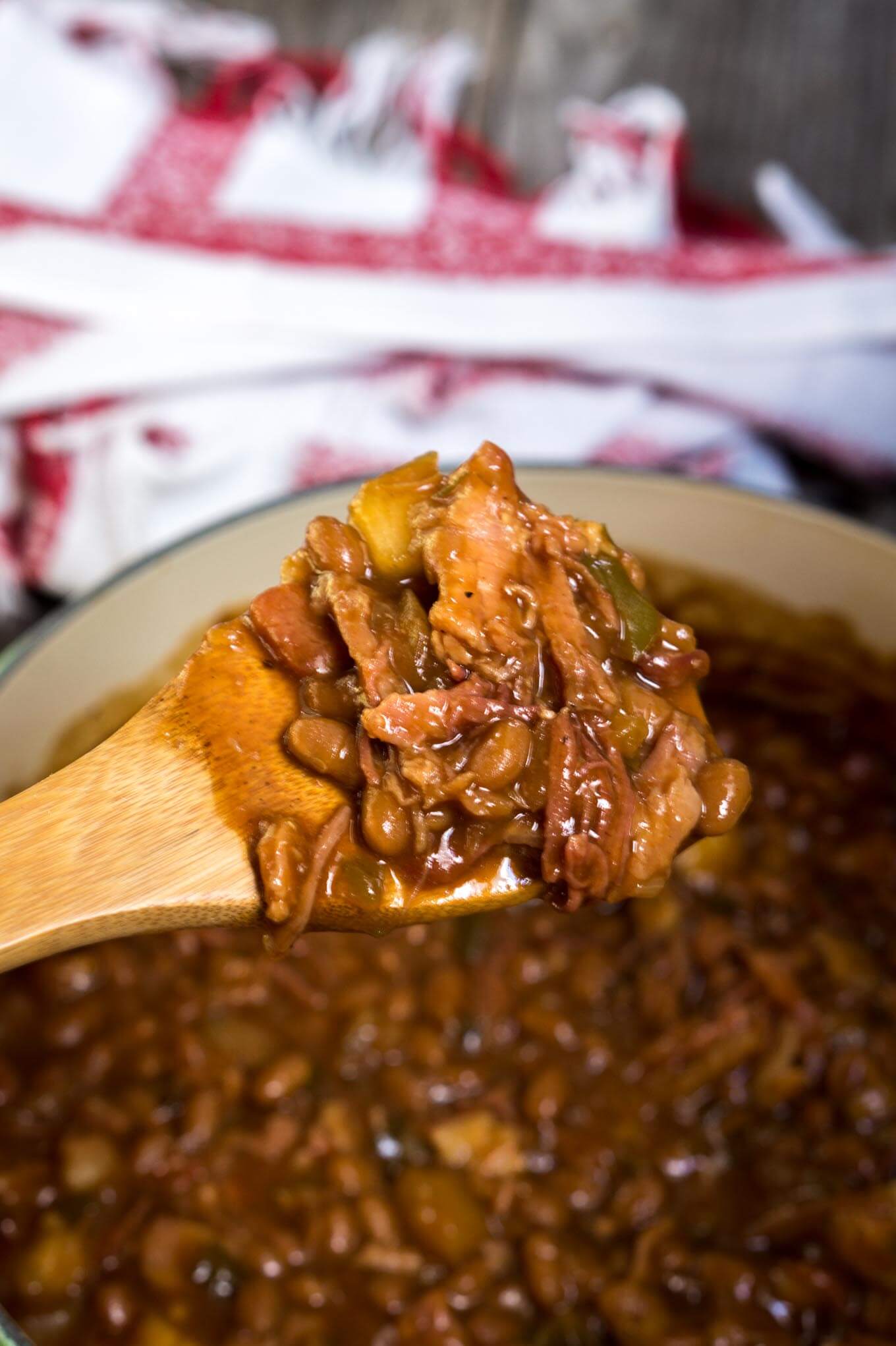 A wooden spoon filled with baked beans, diced apples and shredded pork over a pot filled with Apple Pie Baked Beans