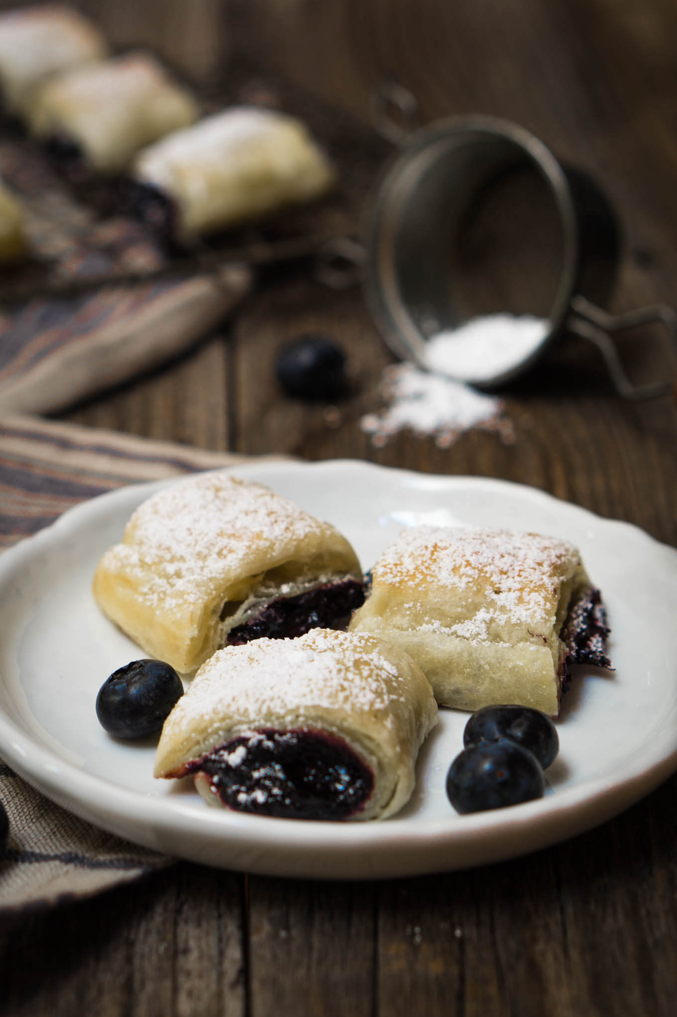 3 blueberry pastry rolls dusted in powdered sugar sitting on a white plate with 3 blueberries on top of a wooden board and in front of spilled powdered sugar.