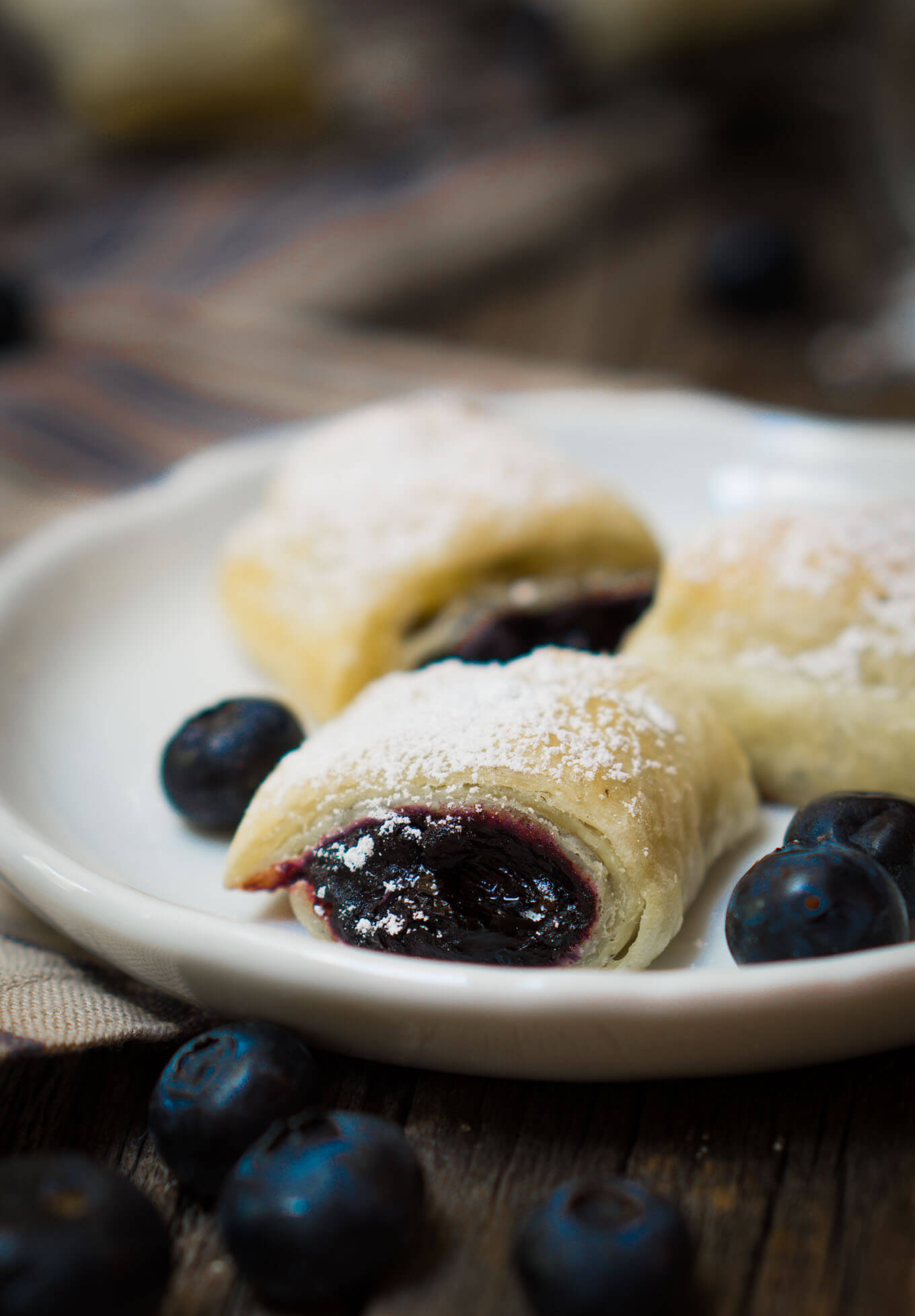  Close up of a blueberry puff pastry roll with bright purple blueberry filling and dusted with powdered sugar sitting on a white plate.