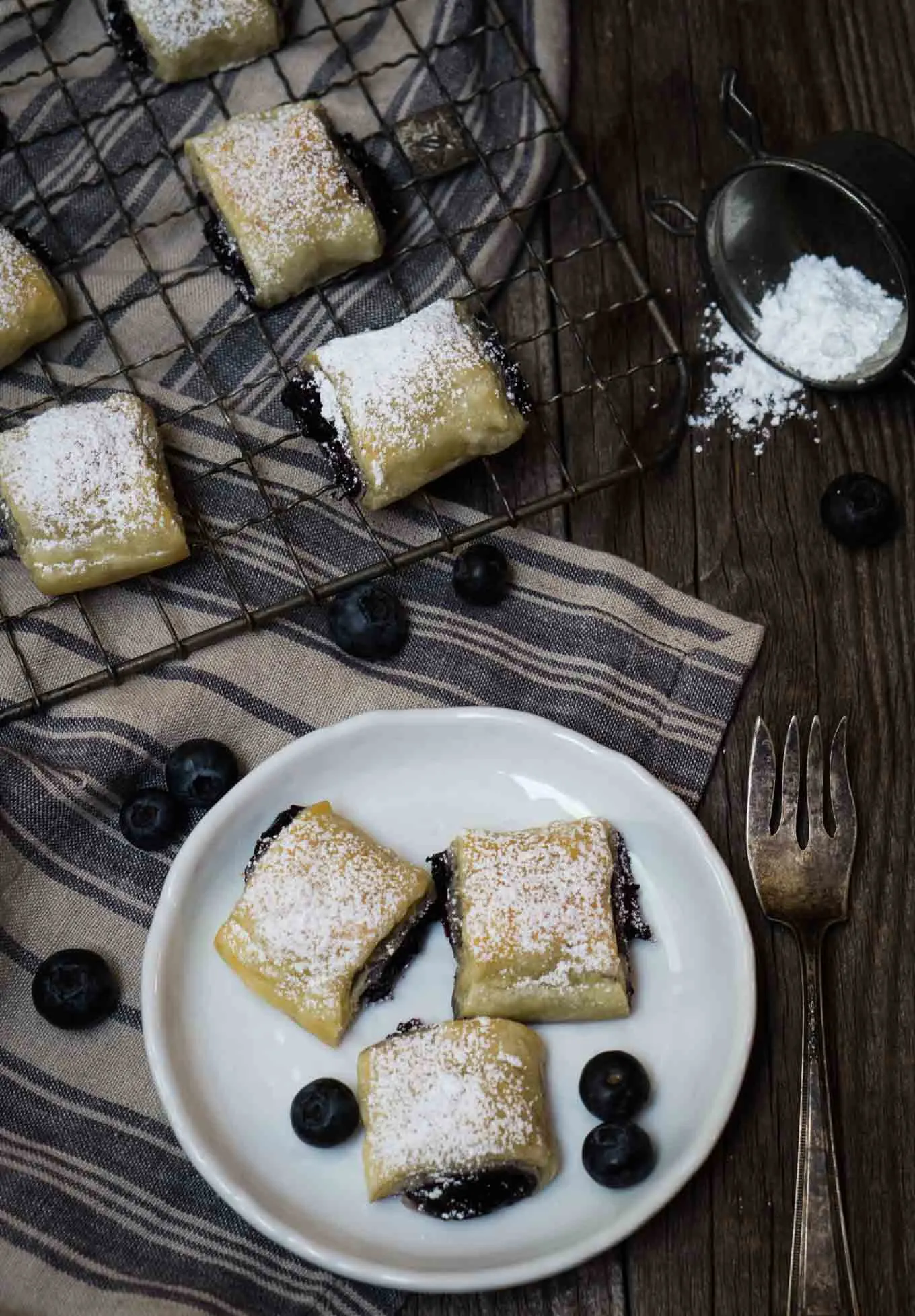 Top shot of 3 blueberry puff pastry rolls sitting on a white plate and more on a cooling rack behind it. Loose blueberries are scattered around. 