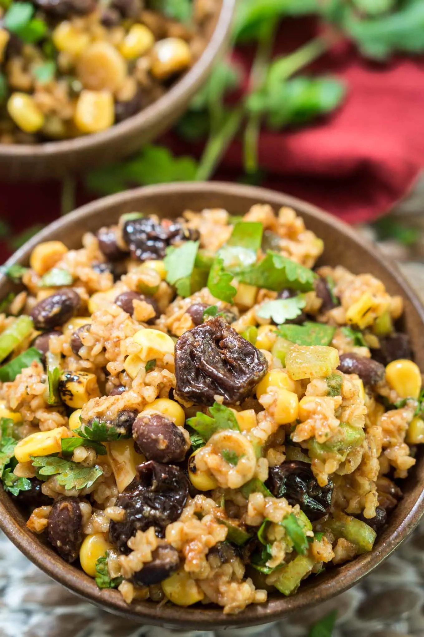 Top-down view of A brown bowl of a sweet and spicy Cherry Chipotle salad loaded with roasted corn, dried cherries, black beans, and garnished with cilantro. A second bowl sits in the background over a red napkin. Fresh cilantro sits to the right of the back bowl.