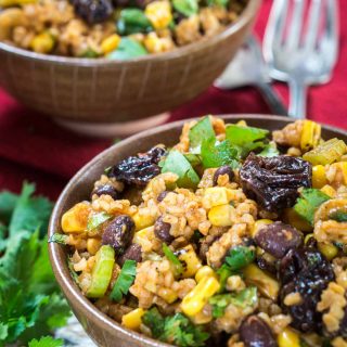 A brown bowl of a sweet and spicy Cherry Chipotle salad loaded with roasted corn, dried cherries, black beans, and garnished with cilantro. A second bowl sits in the background over a red napkin. A fork sits to the right of the back bowl and fresh cilantro to the left of the front bowl.