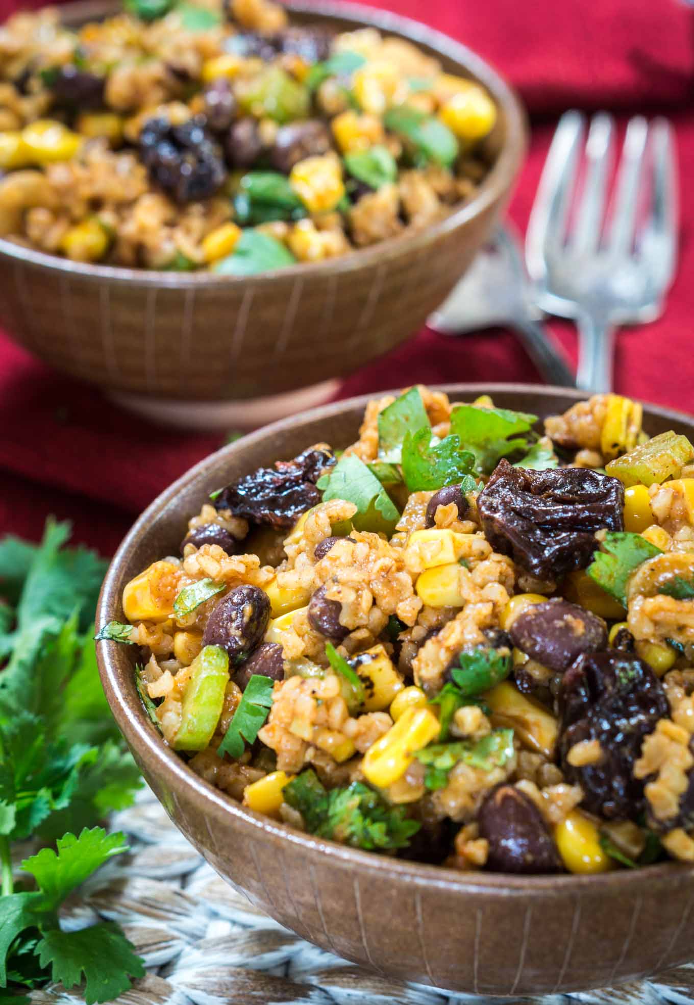 A brown bowl of a sweet and spicy Cherry Chipotle salad loaded with roasted corn, dried cherries, black beans, and garnished with cilantro. A second bowl sits in the background over a red napkin. A fork sits to the right of the back bowl and fresh cilantro to the left of the front bowl.