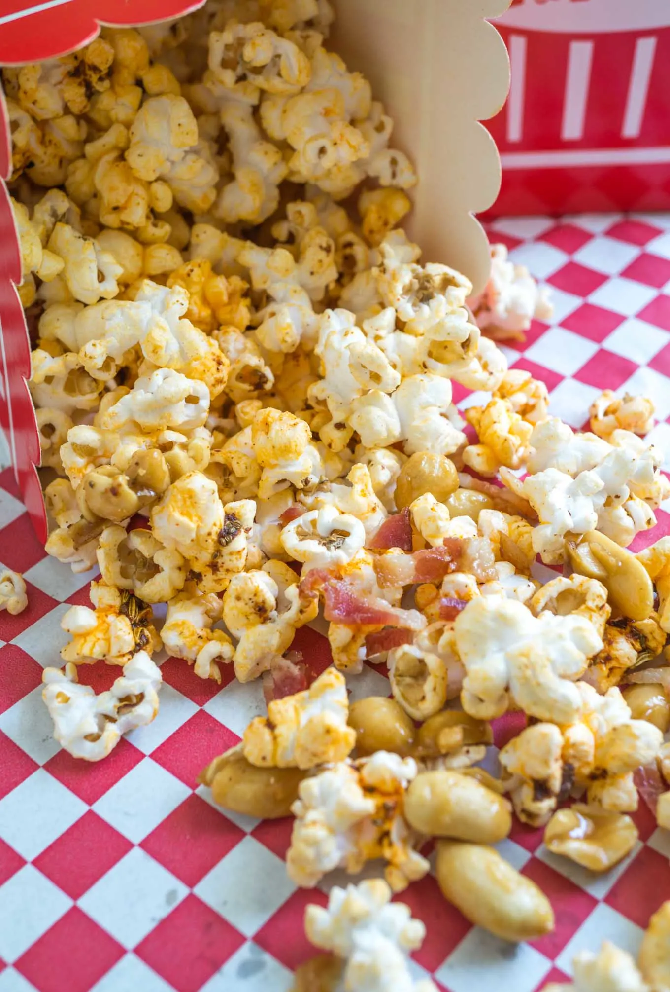 A close-up view of a red and white popcorn box laying on its side with Honey nut popcorn with bacon spilling out onto a red checked napkin.