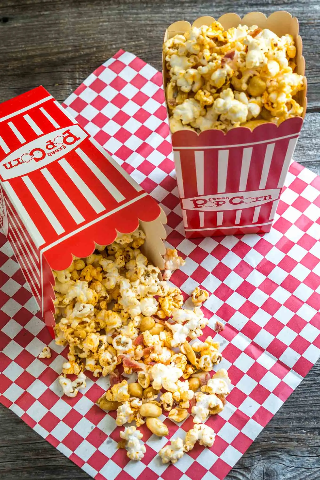 Red stripped popcorn boxes sitting on a red checked napkin. One box is upright and the other on its side with Honey nut popcorn with bacon spilling out.