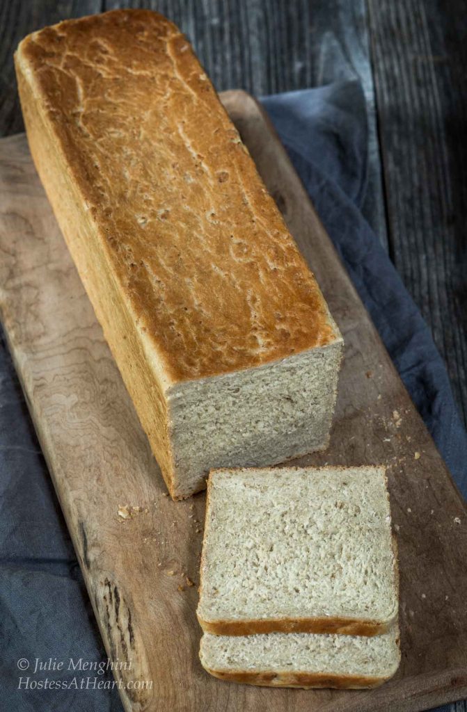 Top-down view of a  loaf of Honey Oat Pain de Mie bread with a slice cut from the front of the loaf sits on a wooden cutting board over a blue napkin.