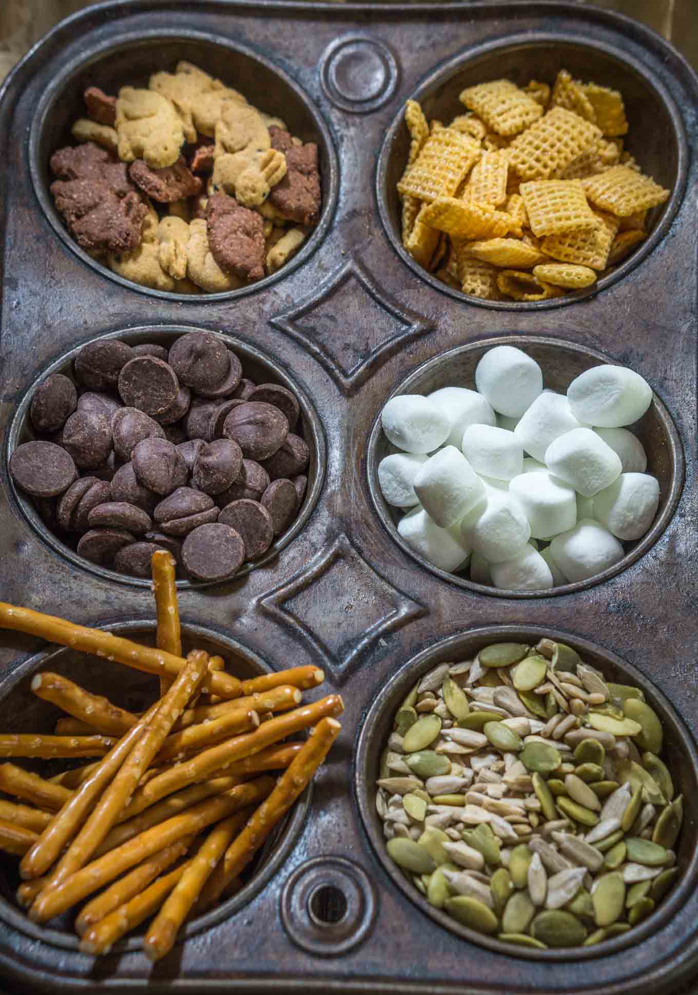 A muffin tin filled with the ingredients for kids trail mix including graham crackers shaped like bunnies, cereal squares, marshmallows, pretzel sticks, and chocolate chips