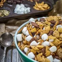 A green bowl filled with snack mix containing graham crackers shaped like bunnies, cereal squares, marshmallows, pretzel sticks, and chocolate chips. A muffin tin is in the back with the ingredients filling the muffin cavities.