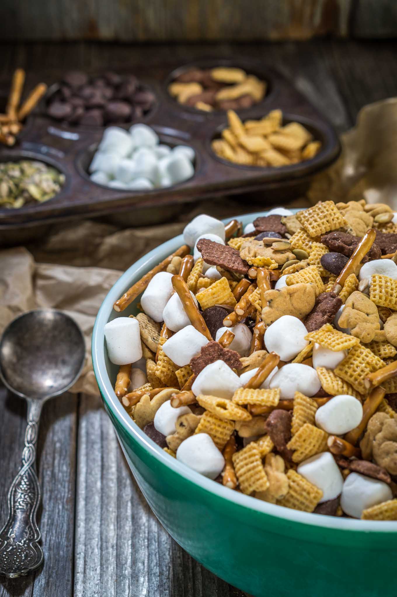 A green bowl filled with snack mix containing graham crackers shaped like bunnies, cereal squares, marshmallows, pretzel sticks, and chocolate chips. A muffin tin is in the back with the ingredients filling the muffin cavities.