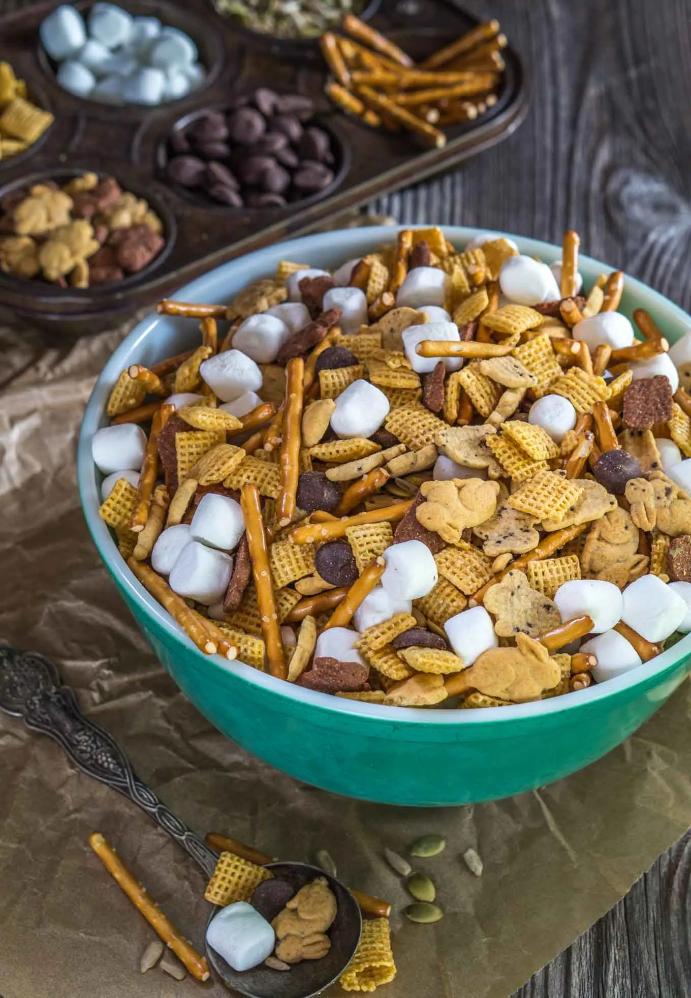 A green bowl filled with snack mix containing graham crackers shaped like bunnies, cereal squares, marshmallows, pretzel sticks, and chocolate chips. A muffin tin is in the back with the ingredients filling the muffin cavities.