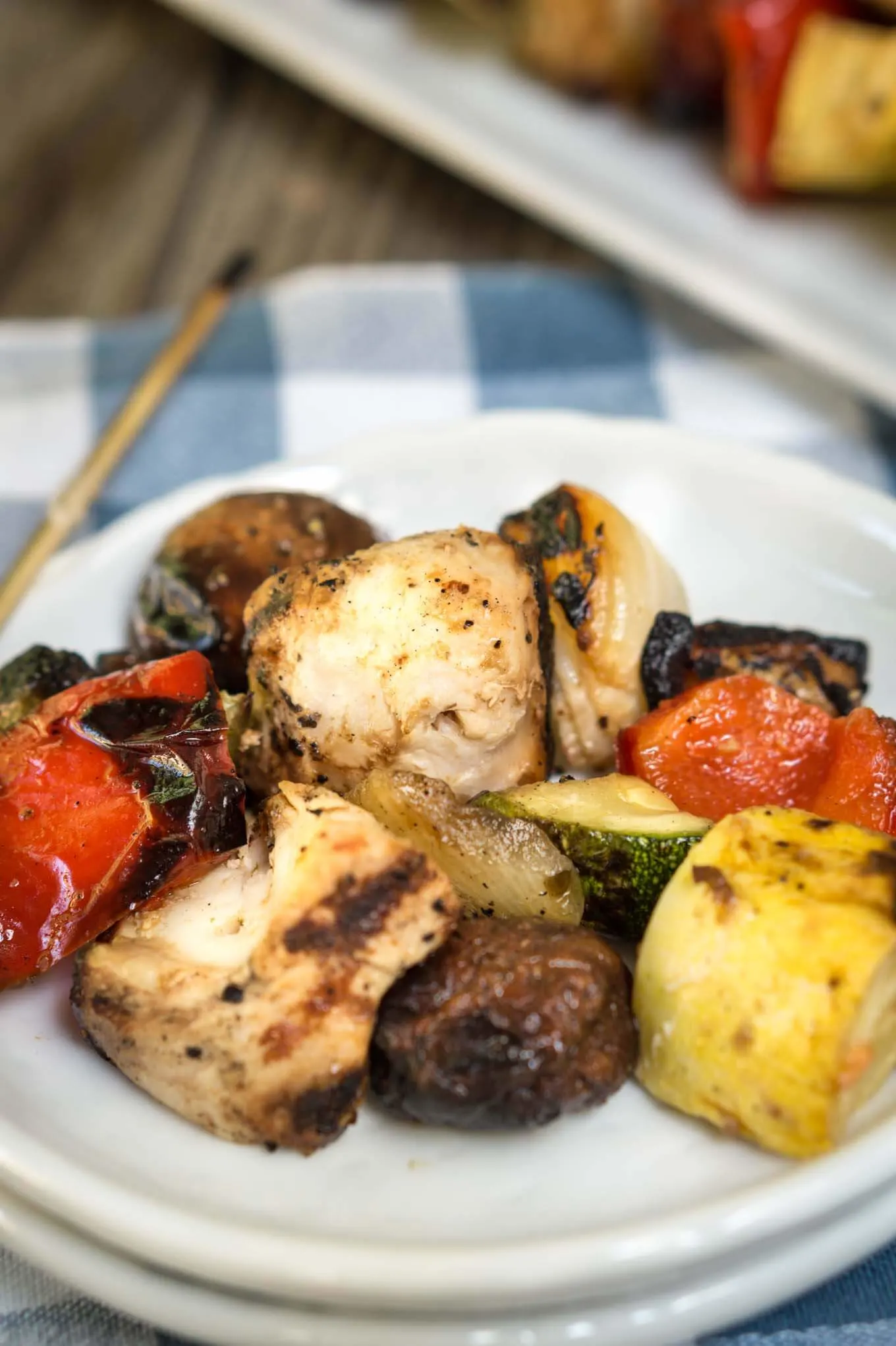 A plate of grilled Shish Kabobs filled with slices of squash, tomatoes, mushrooms, peppers, and chicken on a white plate over a blue checked tablecloth. 