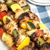 A pile of Shish Kabobs filled with slices of squash, tomatoes, mushrooms, peppers, and chicken on a white plate over a blue checked tablecloth. Empty plates sit in the background.