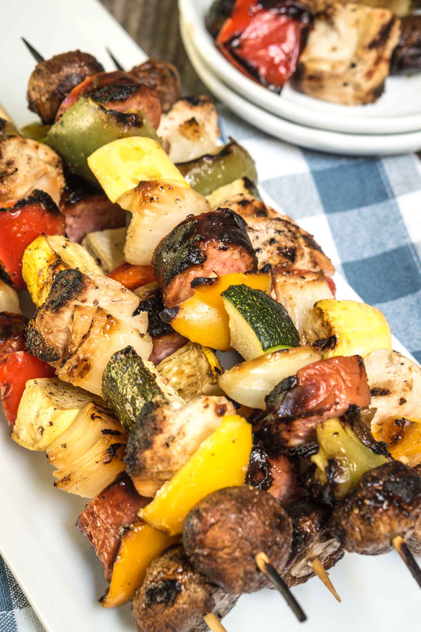 A pile of Shish Kabobs filled with slices of squash, tomatoes, mushrooms, peppers, and chicken on a white plate over a blue checked tablecloth. Empty plates sit in the background.