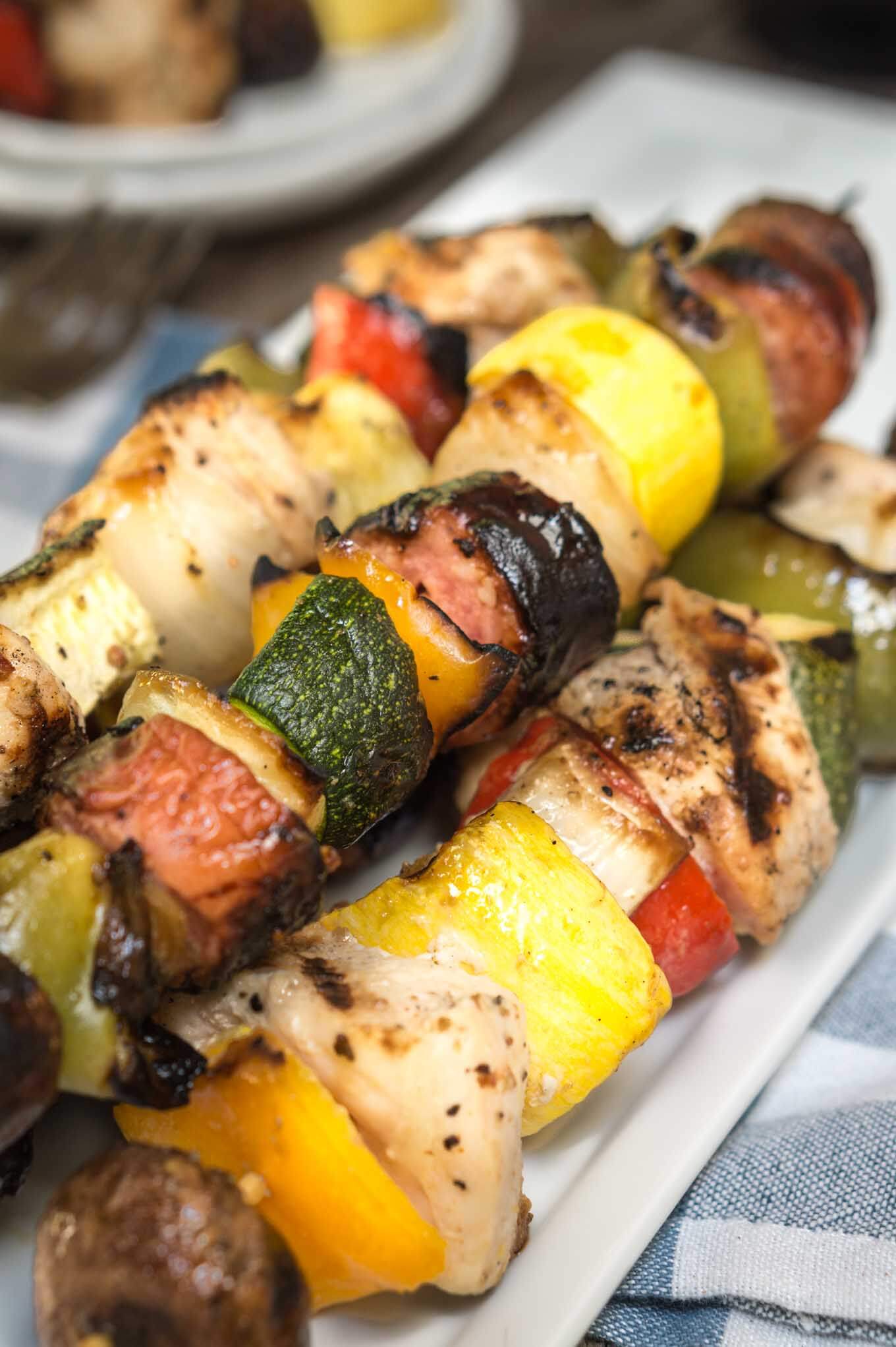 A pile of Shish Kabobs filled with slices of squash, tomatoes, mushrooms, peppers, and chicken on a white plate over a blue checked tablecloth. Empty plates sit in the background.