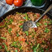 A cast-iron skillet filled with a Sausage and Bulgur Wheat casserole with an antique spoon sitting in it. Fresh tomatoes sit in the background. An antique spoon sits in the center.A pan filled with meat and vegetables, with Bulgur and Wheat