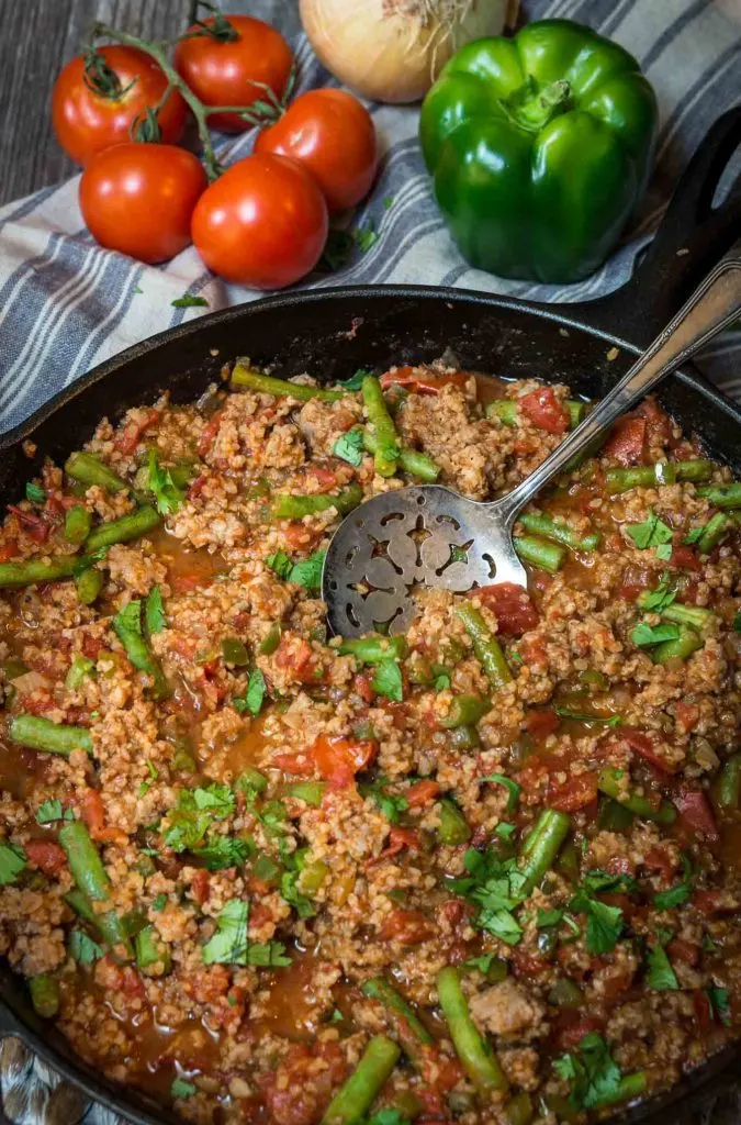 A cast-iron skillet filled with a Sausage and Bulgur Wheat casserole with an antique spoon sitting in it. Fresh tomatoes sit in the background. An antique spoon sits in the center.