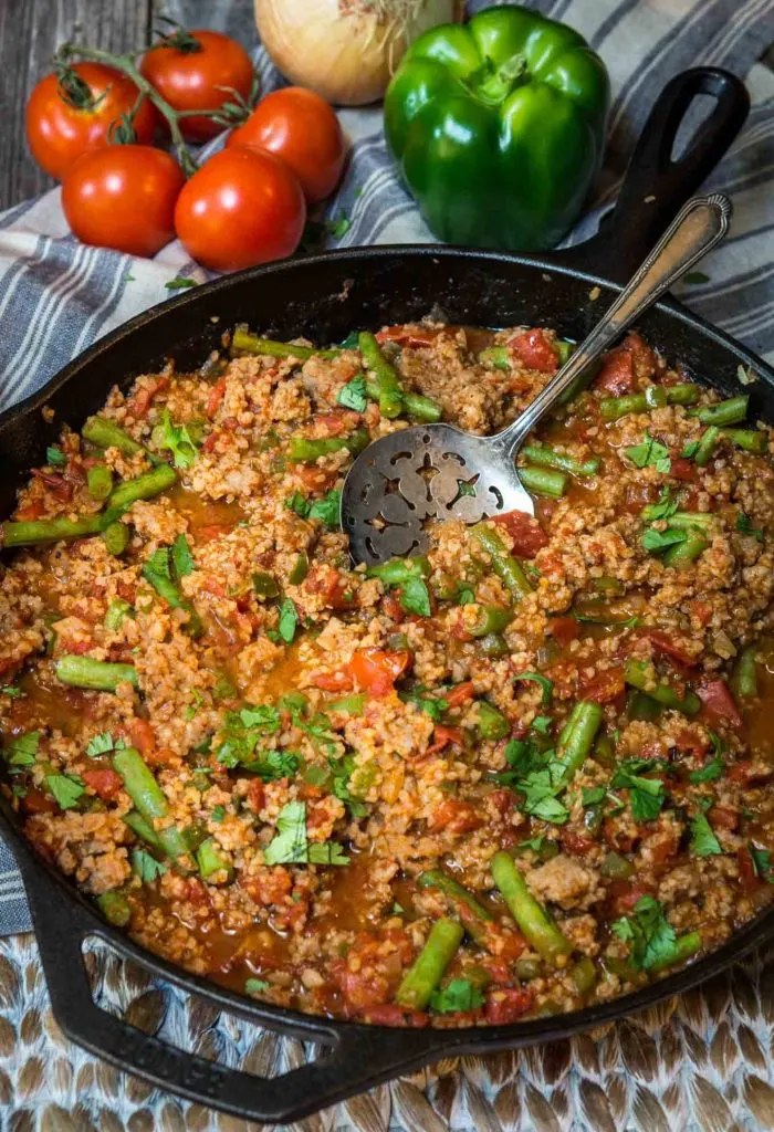 A cast-iron skillet filled with a Sausage Bulger casserole loaded with vegetables and garnished with fresh parsley.