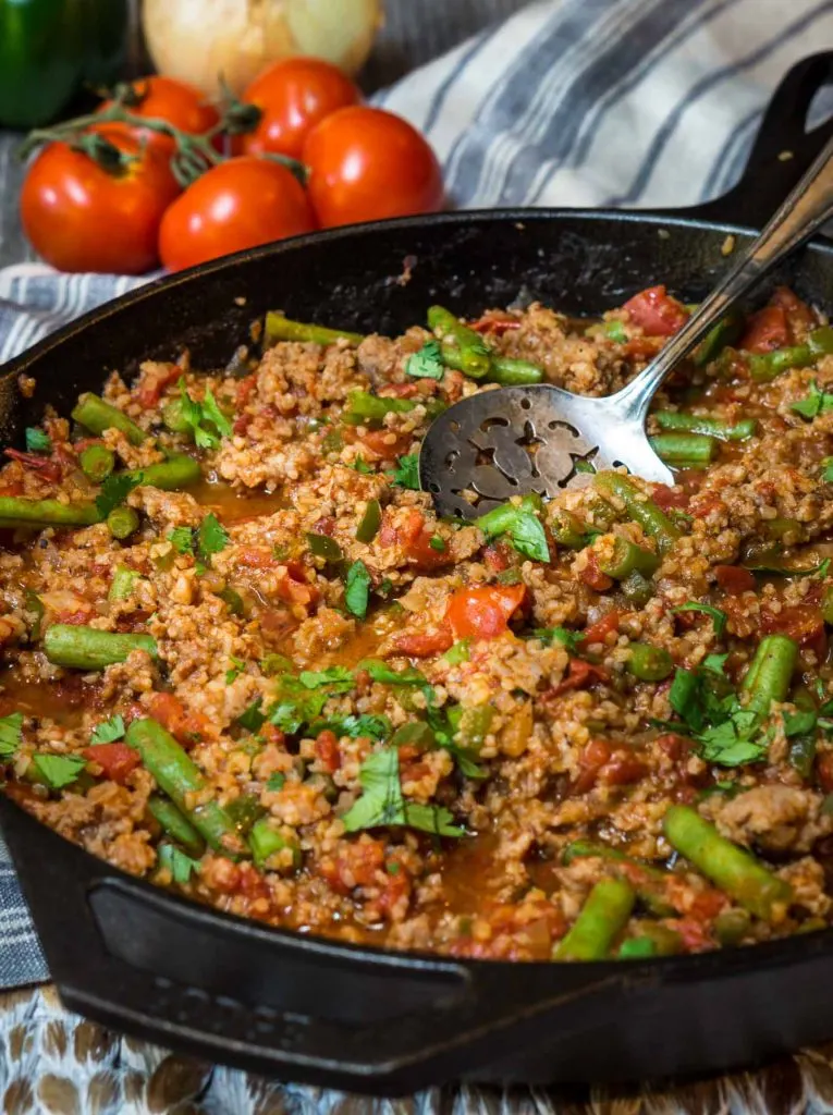A cast-iron skillet full of a casserole filled with Sausage, Bulgur Wheat, Green Beans, and Tomatoes and then garnished with fresh parsley. A large spoon is in the pan. Fresh tomatoes sit in the background over a striped napkin.