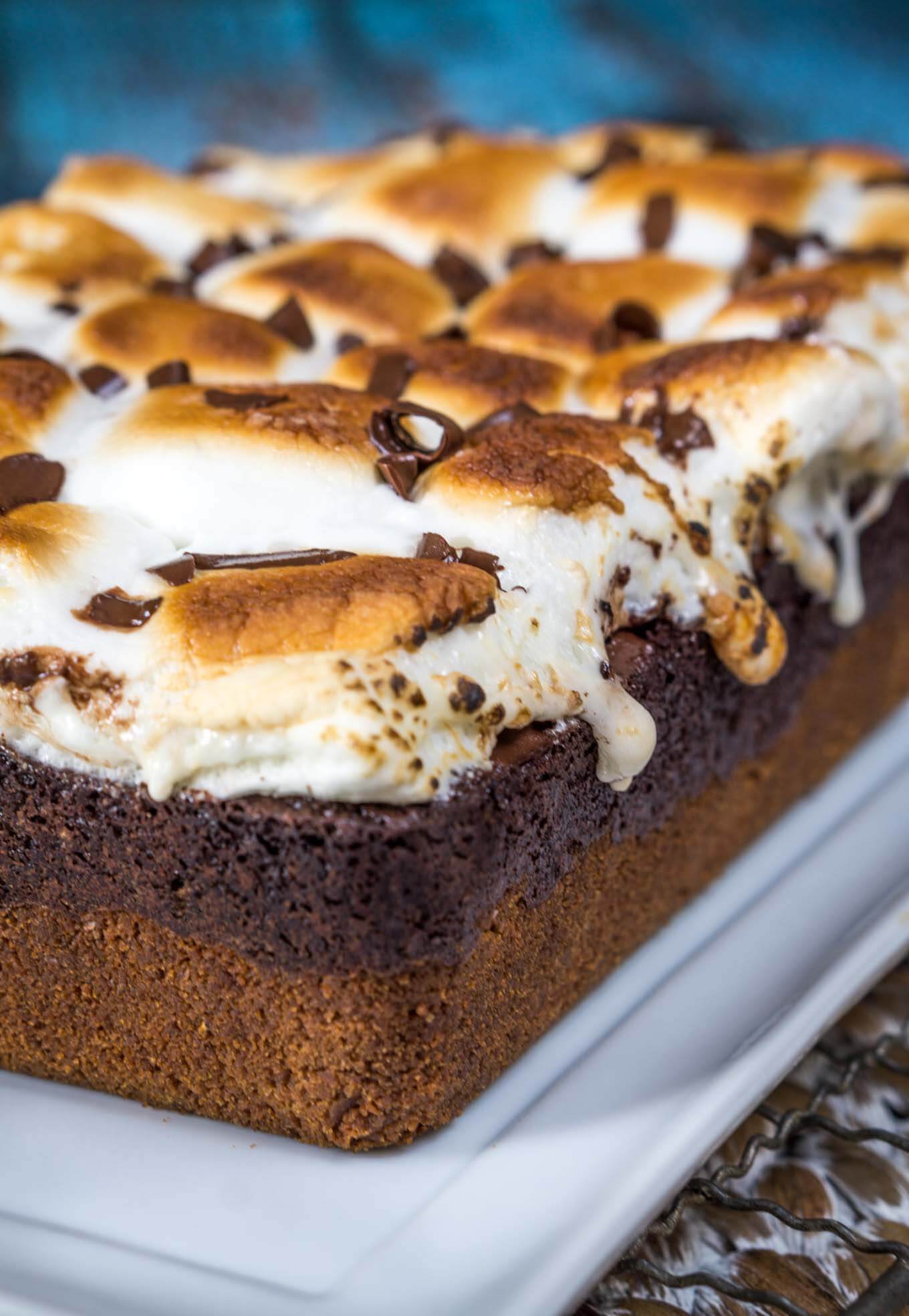 Sideview of a full Smores dessert sitting on a white plate showing the bottom graham cracker crust and topped with a chocolate filling and topped with a toasted marshmallow and chocolate shavings..