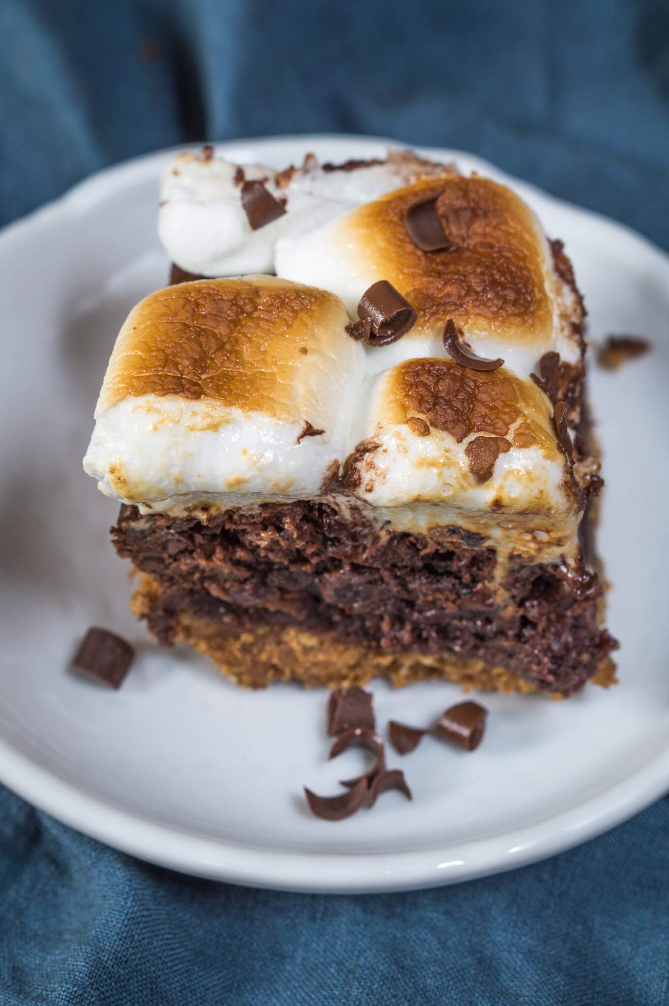 Top down view of a piece of S\'mores chocolate dessert with a bottom layer of graham cracker under a layer of chocolate and topped with toasted marshmallow sitting on a white plate over a blue napkin. A second plate sits in the background.