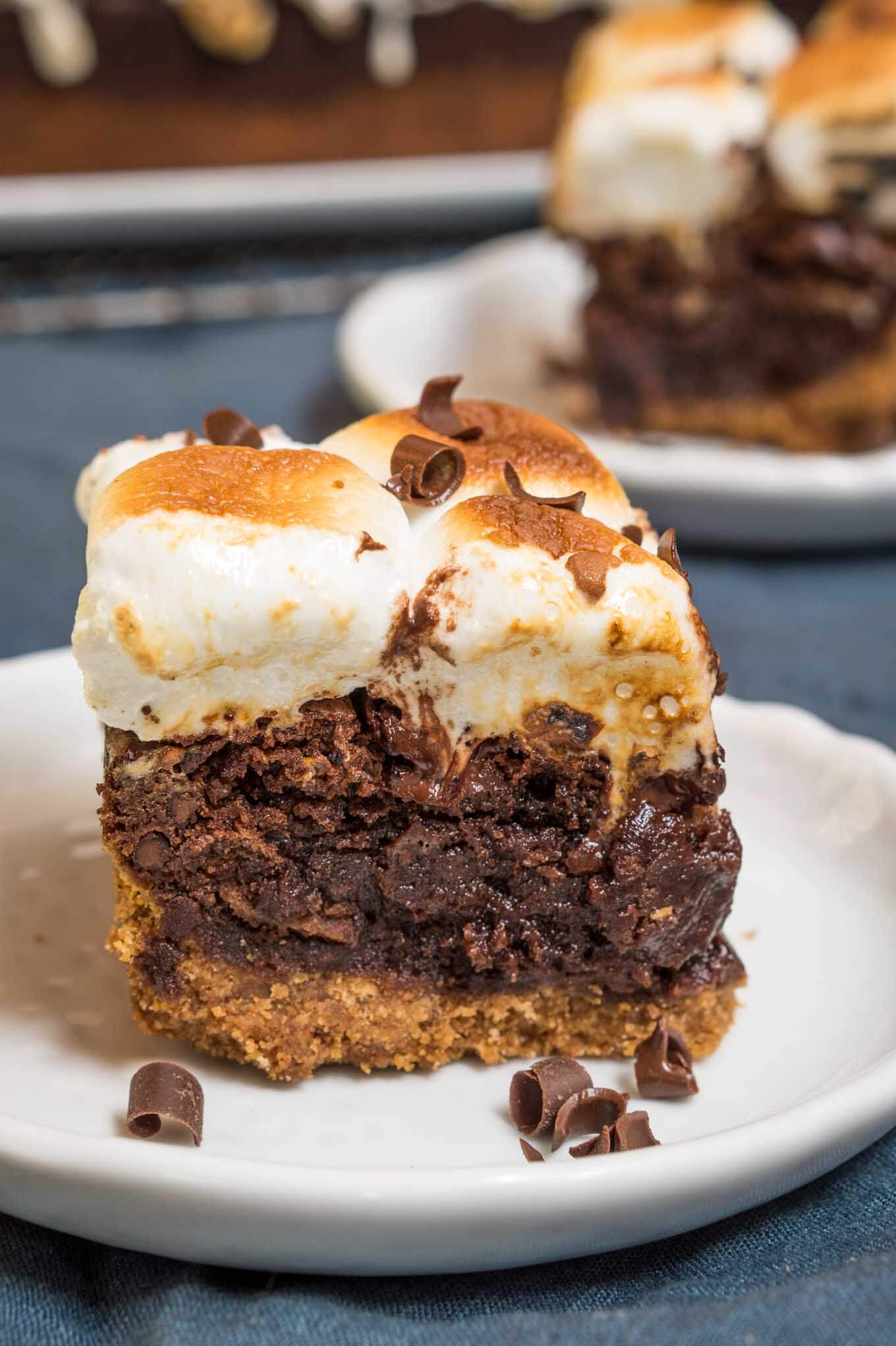 Sideview of a piece of S\'mores chocolate dessert with a bottom layer of graham cracker under a layer of chocolate and topped with toasted marshmallow sitting on a white plate over a blue napkin. A second plate sits in the background.