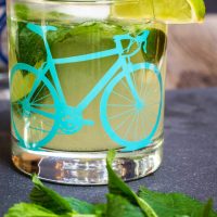 A front view of a slate tray topped with a Mojito topped with fresh mint in a glass with a bicycle on the front. A slice of lime garnishes the glass. Fresh mint sits in the foreground.