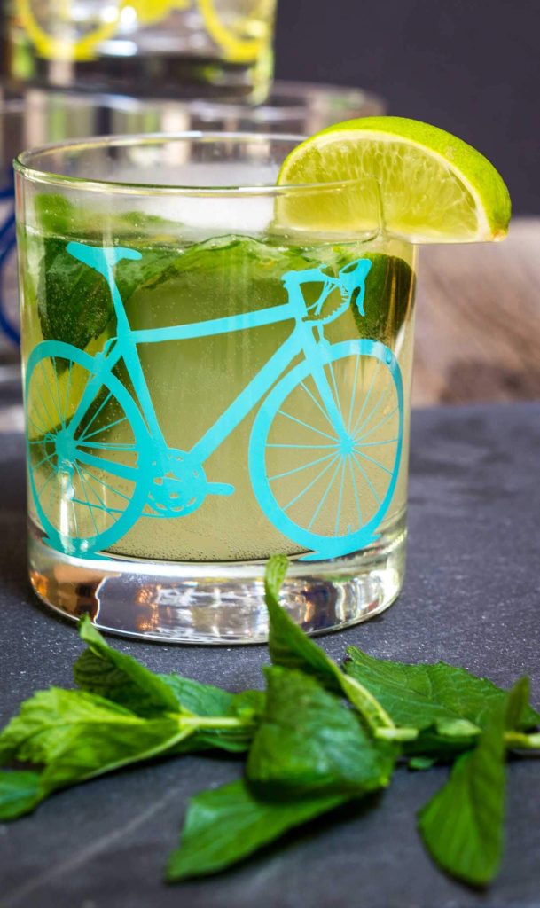 A front view of a slate tray topped with a Mojito topped with fresh mint in a glass with a bicycle on the front. A slice of lime garnishes the glass. Fresh mint sits in the foreground.