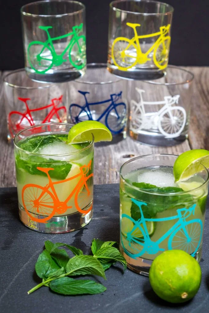 Several glasses with different colored bicycles on the front sitting on a wooden board. Some are filled with mojitos garnished with mint and slices of lime.
