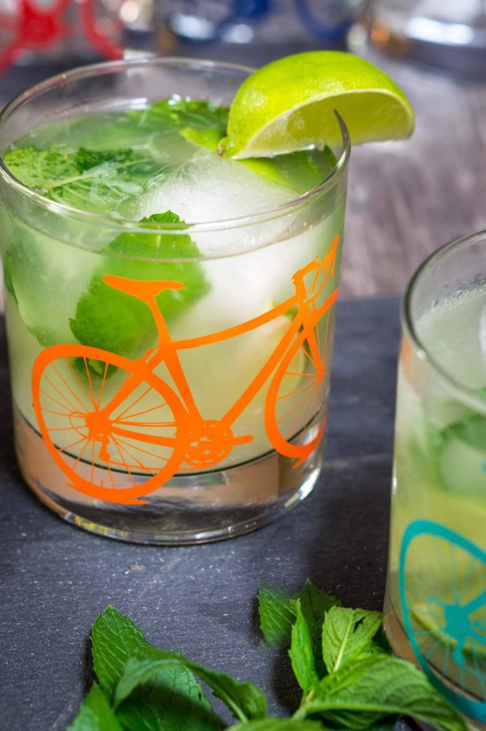 A front view of a slate tray topped with a Mojito topped with fresh mint in a glass with a bicycle on the front. A slice of lime garnishes the glass. A partial glass sits to the right front.