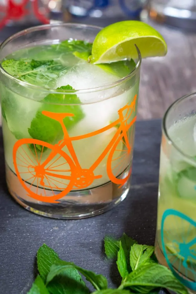 A front view of a slate tray topped with a Mojito topped with fresh mint in a glass with a bicycle on the front. A slice of lime garnishes the glass. A partial glass sits to the right front.