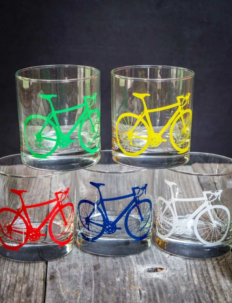 5 glasses with different colored bicycles on the front sitting on a wooden board.