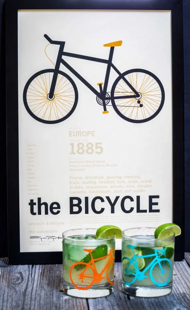 A front view of two Mojitos topped with fresh mint in a glass with a bicycle on the front. A slice of lime garnishes the glass. The glasses sit in front of a print titled \"The Bicycle\".