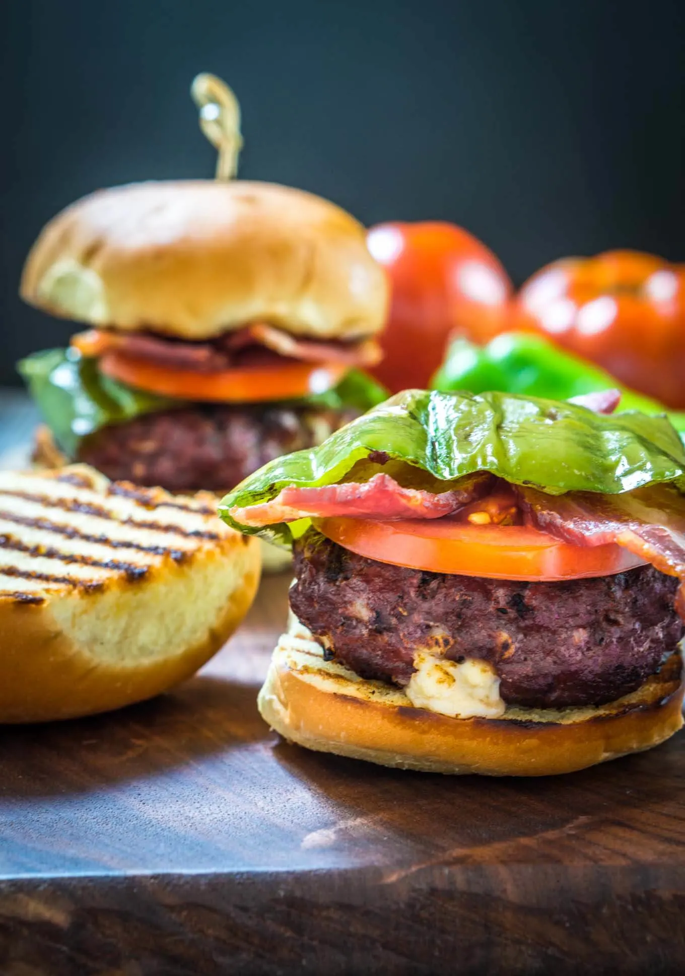 Sideview of a burger stuffed with hatch chile and Gruyere layered with lettuce, tomato, a hatch chile, and bacon with the top of the toasted bun sitting next to it. A second burger sits in the background next to fresh tomatoes.