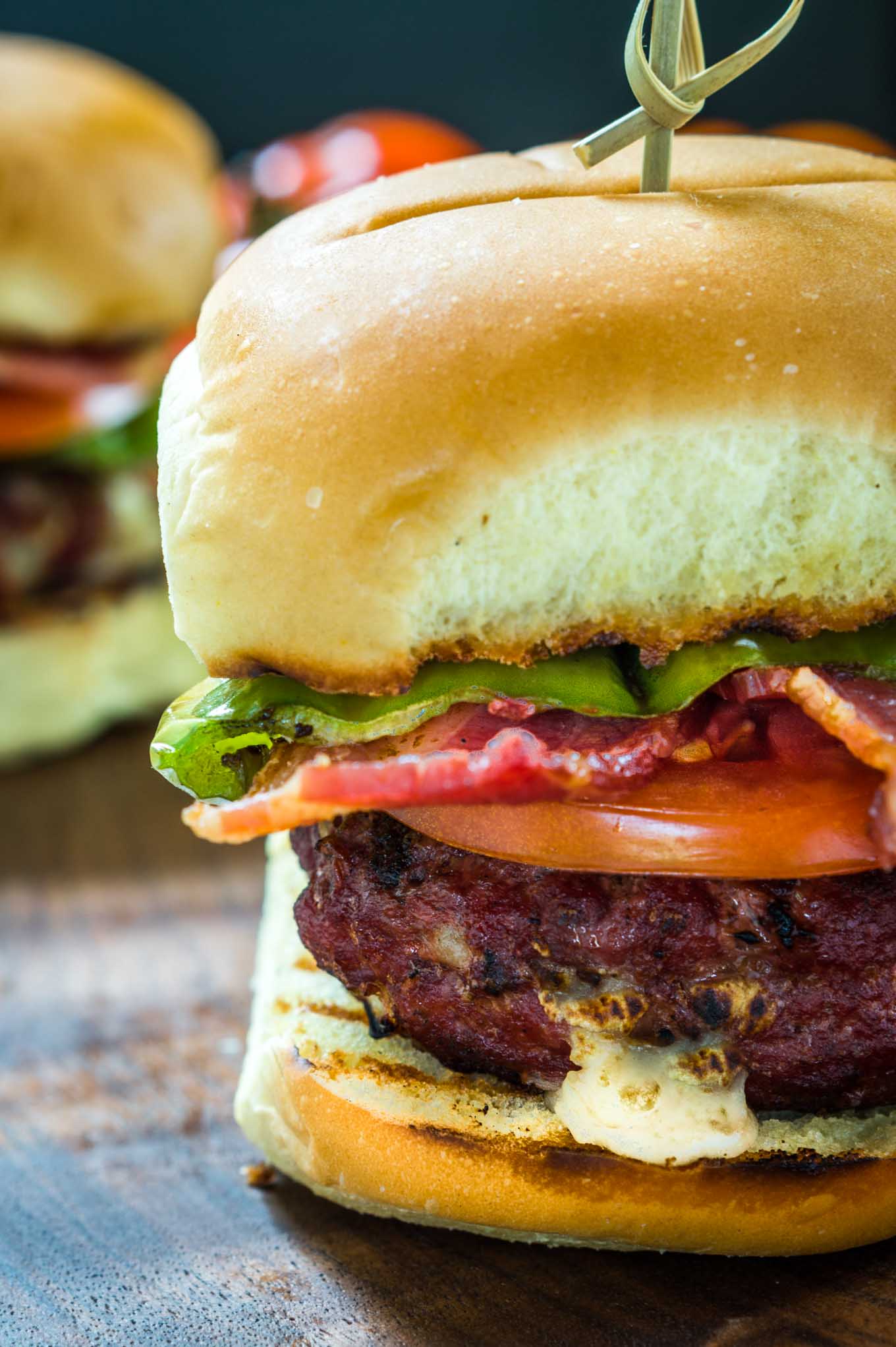 Sideview close-up of a burger stuffed with hatch chile and Gruyere layered with lettuce, tomato, a hatch chile, and bacon secured with a bamboo skewer. A second burger sits in the background.