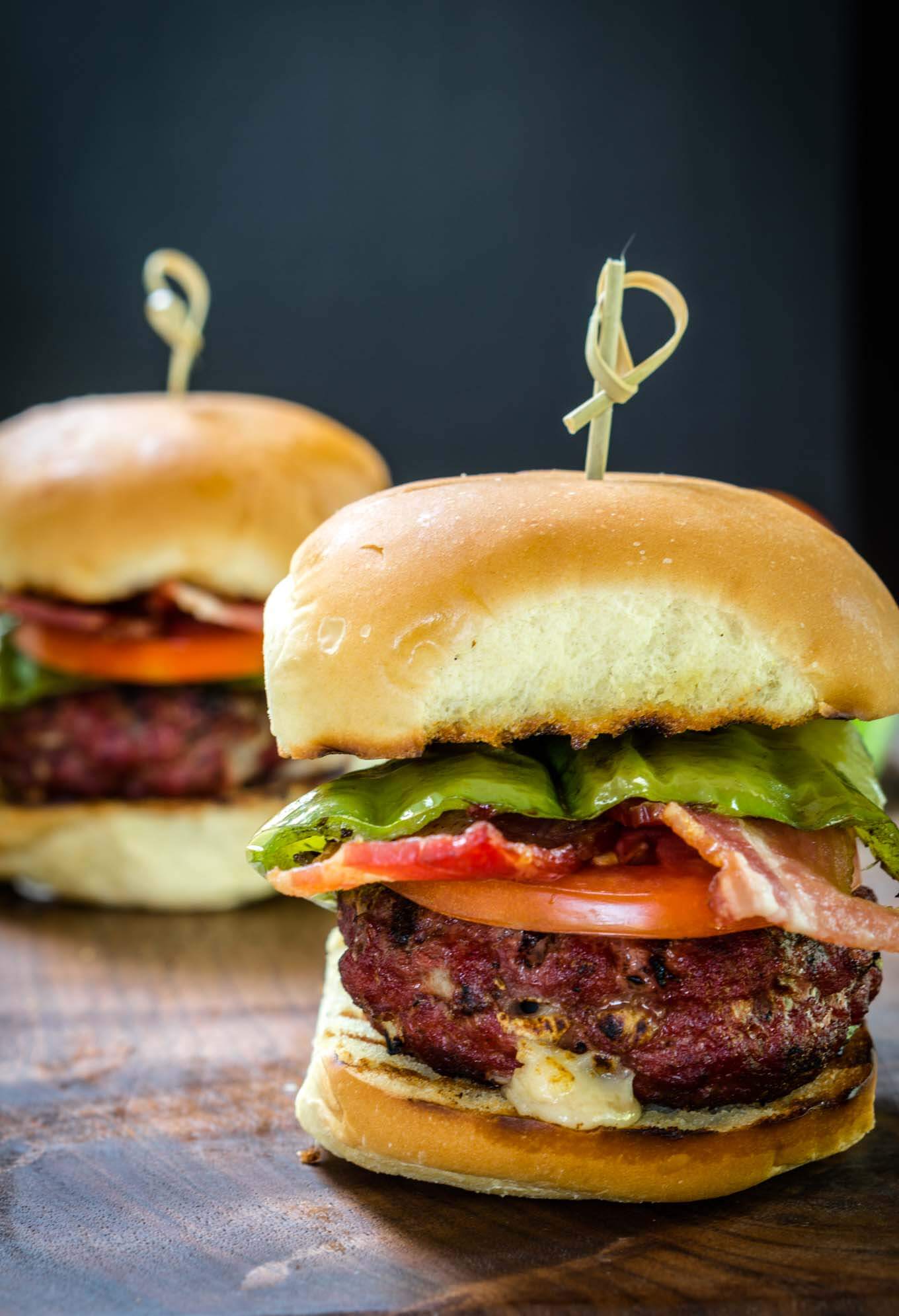 Sideview of a burger stuffed with hatch chile and Gruyere layered with lettuce, tomato, a hatch chile, and bacon secured with a bamboo skewer. A second burger sits in the background.