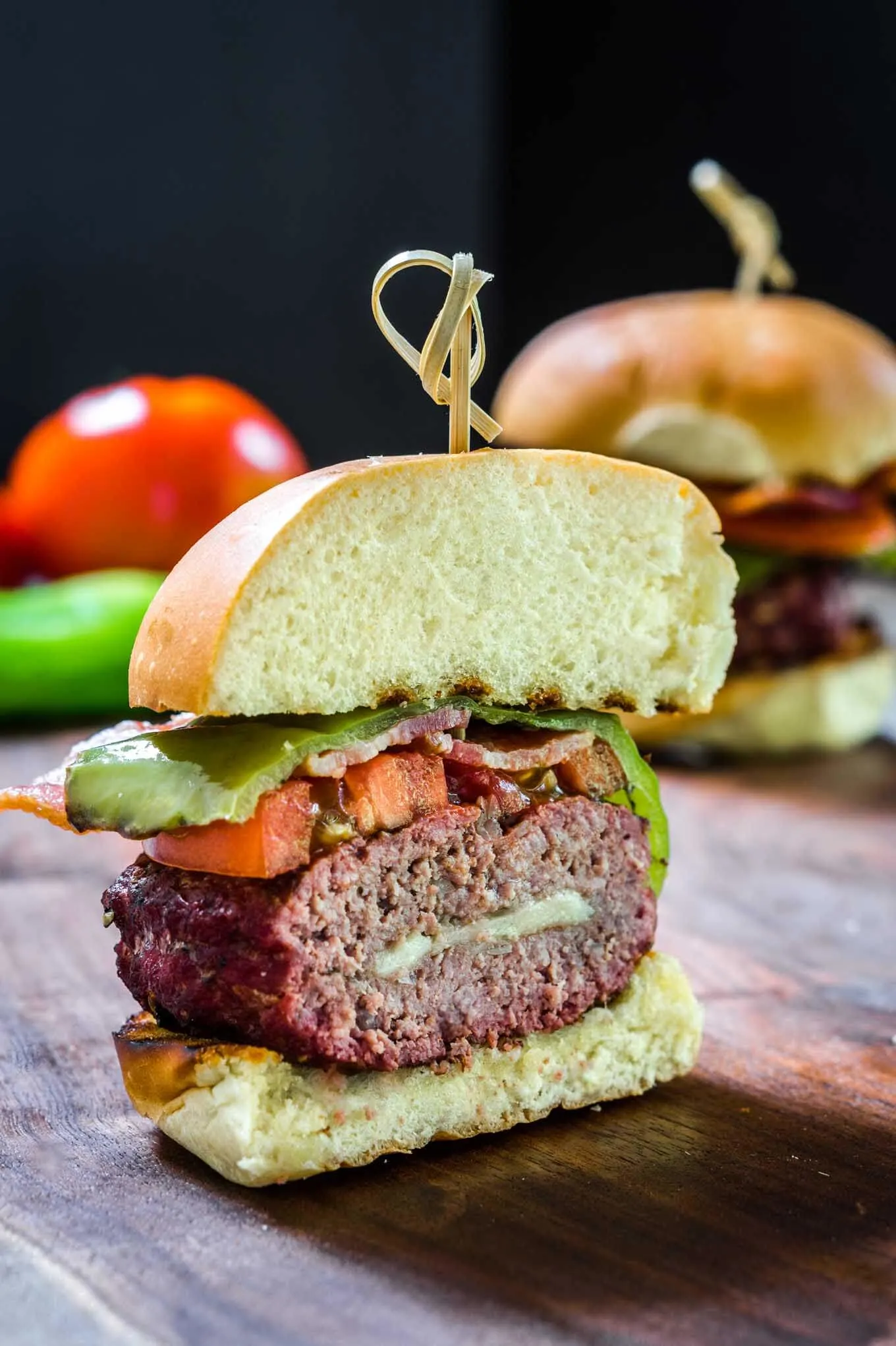  A Hatch Chile Gruyere Burger on a bun cut in half showing how its stuffed with cheese and chile and layered with bacon, tomato, and chile then secured with a bamboo skewer.