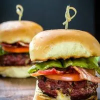 Side view of a hamburger that's been stuffed with Pepperjack cheese and pepperoncini sitting over a slice of tomato and lettuce between a bun. A skewer secures the bun to the burger. A second burger sits in the background.