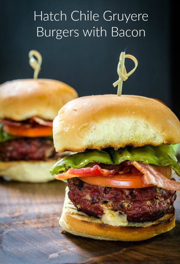 Sideview of a burger stuffed with hatch chile and Gruyere layered with lettuce, tomato, a hatch chile, and bacon secured with a bamboo skewer. A second burger sits in the background.