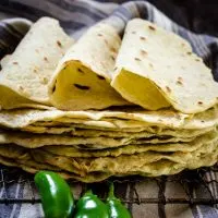 A stack of homemade tortilla shells on a cooling rack with three folded tortillas on top. 3 fresh jalapenos sit in the front and a blue striped towel sits in the background.