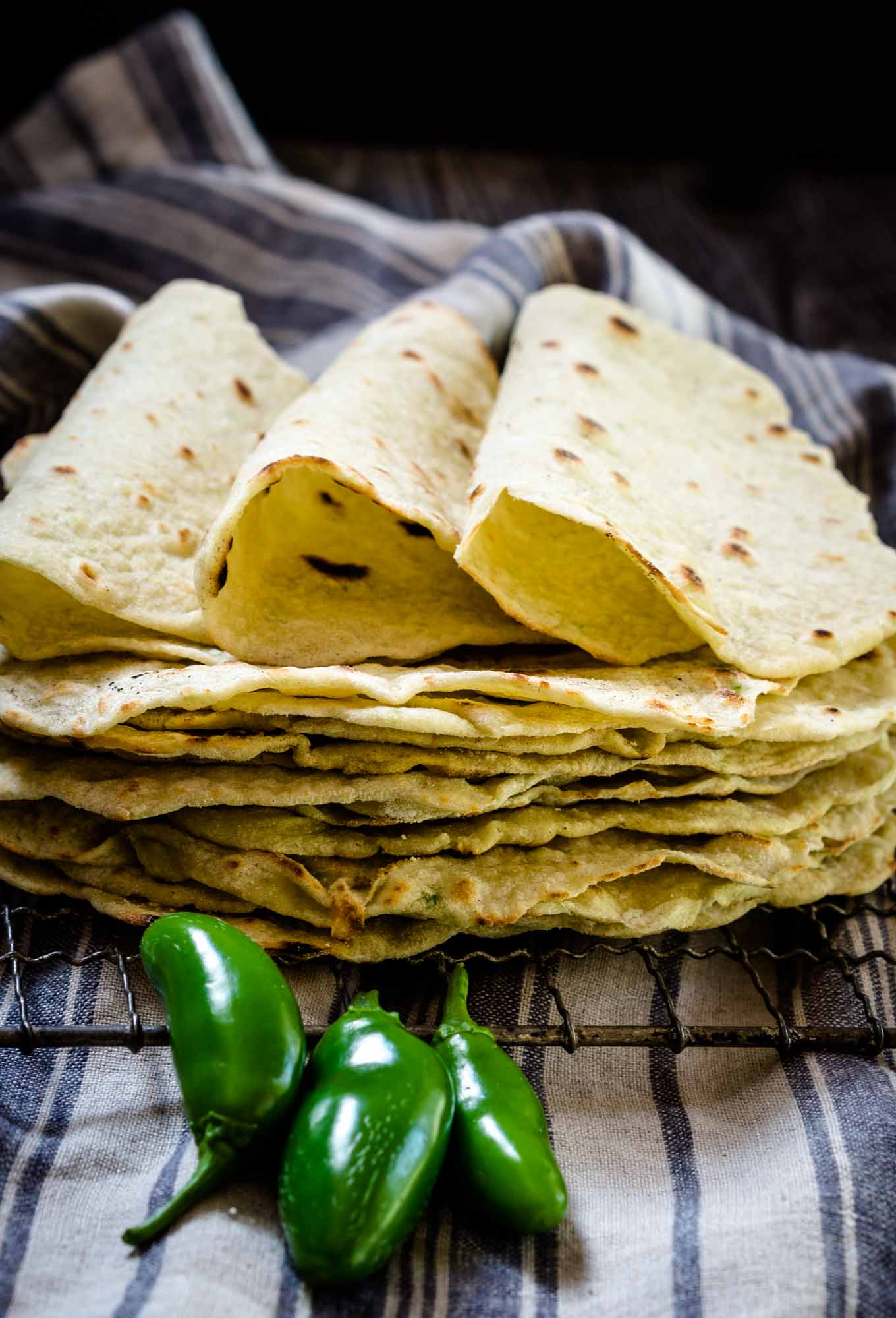 A stack of homemade tortilla shells on a cooling rack with three folded tortillas on top. 3 fresh jalapenos sit in the front and a blue striped towel sits in the background.