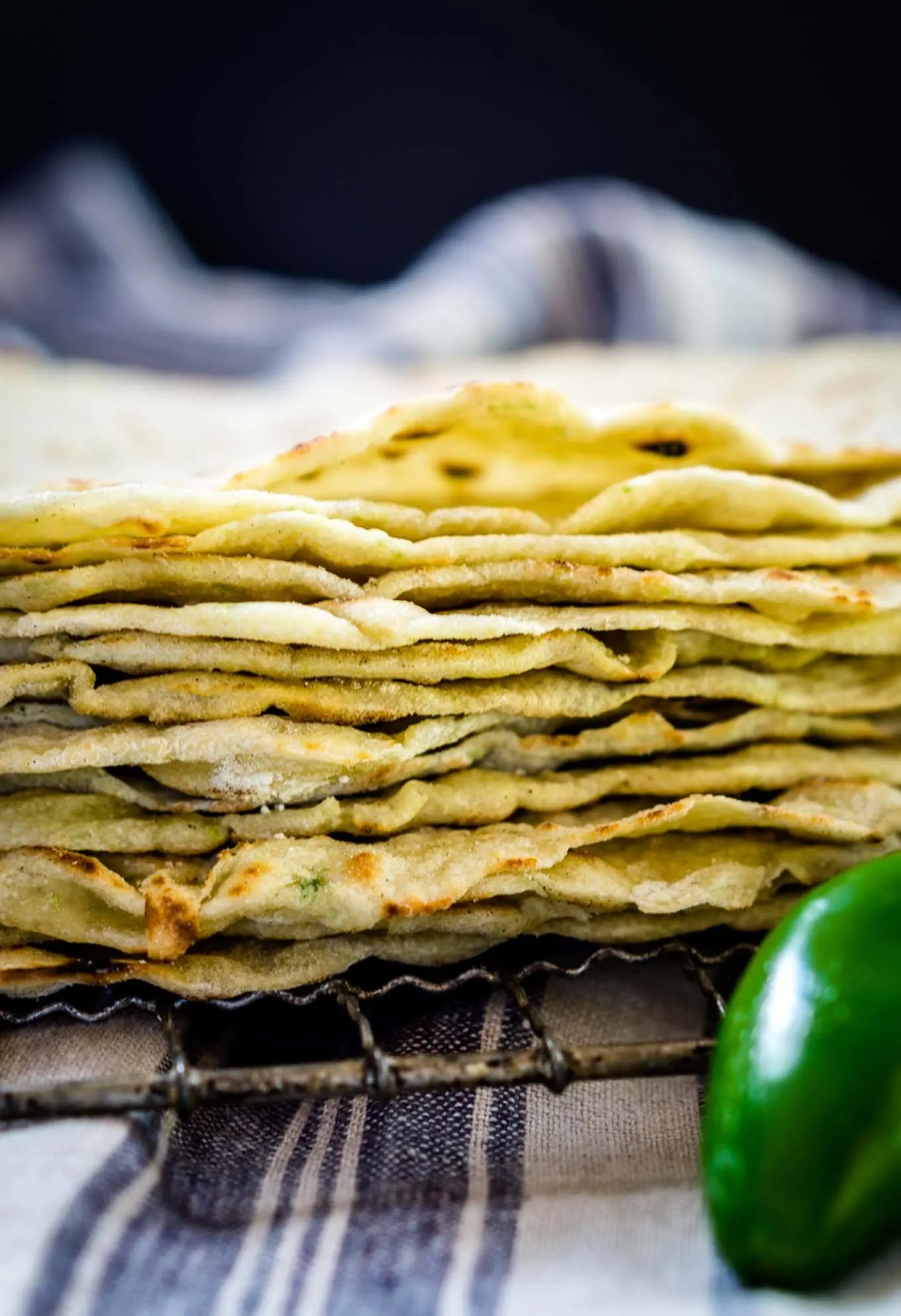 A stack of homemade tortilla shells on a cooling rack. A fresh jalapeno sits in the front and a blue striped towel sits in the background.